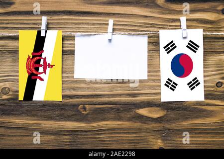 Hanging flags of Brunei and South Korea attached to rope with clothes pins with copy space on white note paper on wooden background.Diplomatic relatio Stock Photo