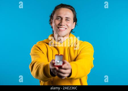 Young handsome guy holds out jewelry box with wedding ring to the camera. Guy makes marriage proposal. He is smiling. Stock Photo