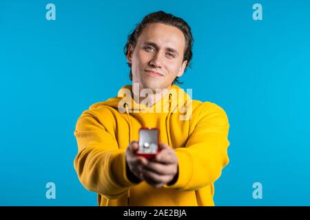 Young handsome guy holds out jewelry box with wedding ring to the camera. Guy makes marriage proposal. He is smiling. Stock Photo