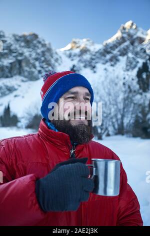 Traveler man with beard is drinking tea or coffee from cup in mountains at winter time. Mountain camping concept. Stock Photo