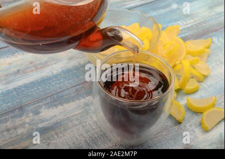 Black tea is poured from a teapot into a glass, marmalade lemon slices on a wooden background. Close up Stock Photo