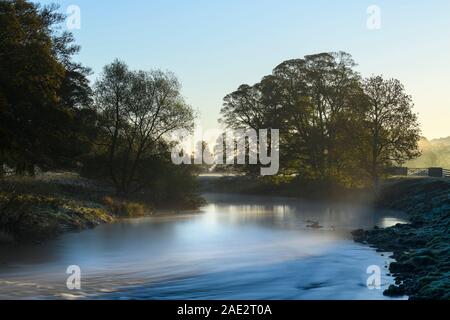 Cold early morning sunrise in scenic countryside, mist or fog lying over water of River Wharfe - Burley in Wharfedale, West Yorkshire, England, UK. Stock Photo