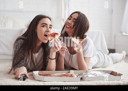 Just having fun. Sisters eating pizza when watching TV while lying on the floor of beautiful bedroom at daytime Stock Photo