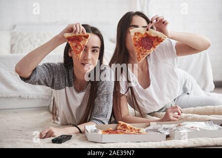 Holds food in the hands. Sisters eating pizza when watching TV while lying on the floor of beautiful bedroom at daytime Stock Photo