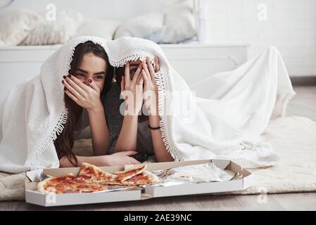 One girl is smiling, other is scared. Warm blanket. Sisters eating pizza when watching TV while lying on the floor of beautiful bedroom at daytime Stock Photo