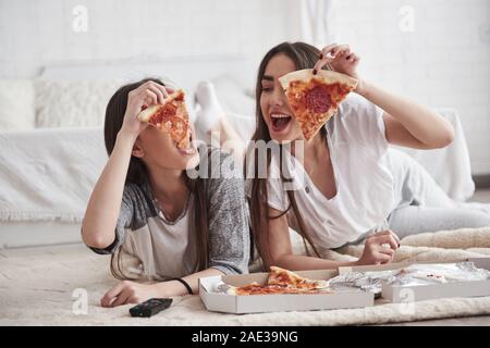 Delicious slices. Sisters eating pizza when watching TV while lying on the floor of beautiful bedroom at daytime Stock Photo