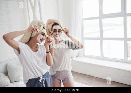 Ordinary day of a girls. Conception of skin care by using white mask and cucumbers on the face. Two female sisters have weekend at bedroom Stock Photo