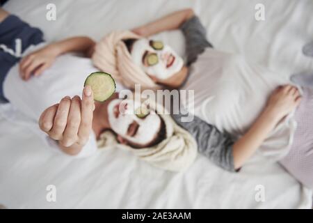 Conception of self care. Girl holds cucumber ring in the hand. Lying on the white bed. Top view Stock Photo