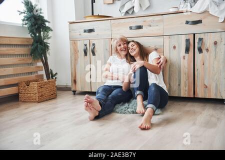 Smiling together when sitting on the floor. Mother and daughter having good time in the kitchen Stock Photo
