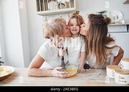 Kissing lovely child from two sides. Mother, grandmother and daughter having good time in the kitchen