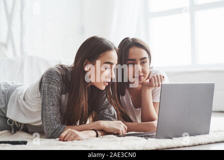 Surfing in social medias. Young female twins lying on the floor near white bed and using silver colored laptop Stock Photo