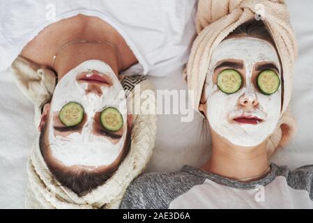 Lying upside down. Conception of skin care by using fresh cucumber rings and white mask on the face. Two female sisters have weekend at bedroom Stock Photo