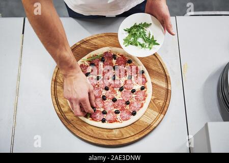 Work in progress. Top view of baker in white shirt putting olives to make delicious pizza for an order in restaurant Stock Photo