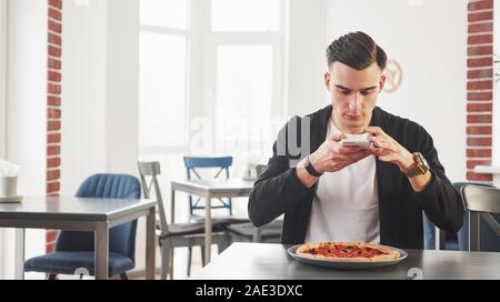 Takes photo using smartphone camera. Having a breakfest with pizza at daytime. Young stylish man in black jacket Stock Photo