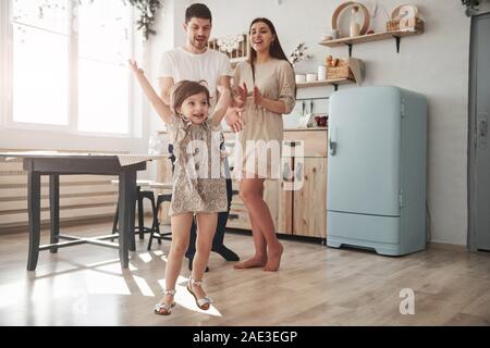 With hands up. Playful female child have fun by running in the kitchen at daytime of front of her mother and father Stock Photo