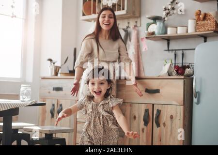 Be happy like this little girl. Playful female child have fun by running in the kitchen at daytime of front of her mother Stock Photo