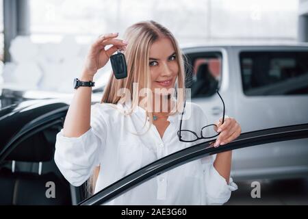 With keys in hand. Cute girl in eyewear stands near the car in auto saloon. Probably her next purchase Stock Photo