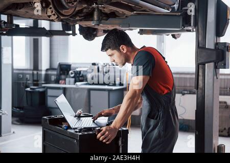 Black hair. Man at the workshop in uniform using laptop for his job for fixing broken car Stock Photo