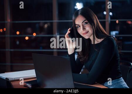 Helps on the phone even when it's that late. Attractive brunette businesswoman works alone in the office at nightime Stock Photo