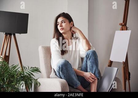 Relaxed woman sitting on the chair and looking to the side. Young brunette in the room with white walls and daylight that comes from the window Stock Photo