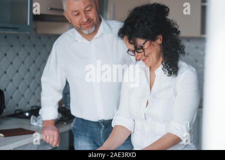 Having good mood. Man and his wife in white shirt preparing food on the kitchen using vegetables Stock Photo