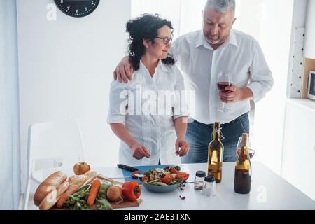 Doing work together. Man and his wife in white shirt preparing food on the kitchen using vegetables Stock Photo