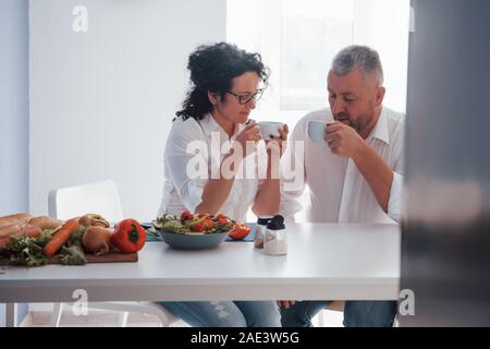 Calm time spending. Senior man and his wife in white shirt have romantic dinner on the kitchen Stock Photo