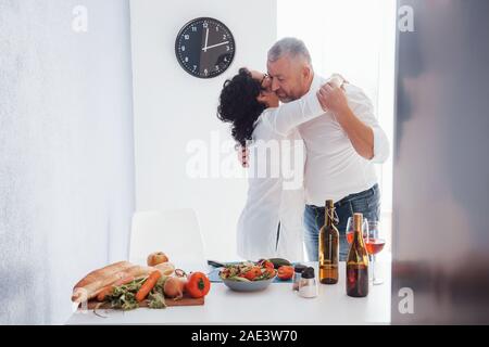 Time for embracing and dancing. Senior man and his wife in white shirt have romantic dinner on the kitchen Stock Photo