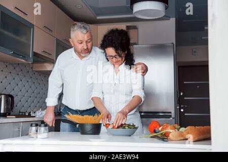 Good looking people. Man and his wife in white shirt preparing food on the kitchen using vegetables Stock Photo