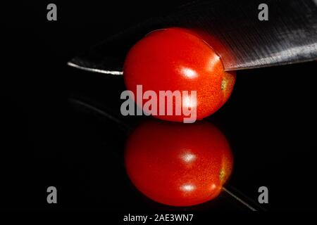 A single grape tomato on a highly reflective surface being sliced by a knife on a black background with vibrant colors and high detail. Stock Photo