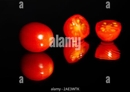 Grape tomatoes against a black background being sliced with a sharp knife on a highly reflective surface.  Vibrant colors Stock Photo