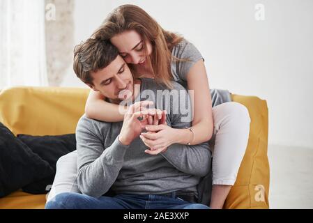 Girl hugs man from behind. Happy couple relaxing on the yellow sofa in the living room of their new house Stock Photo