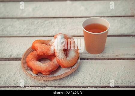 Russian fast food bagel pyshki and paper cup with tea or coffe on wooden table, covered with snow, winter snack in the park. Stock Photo