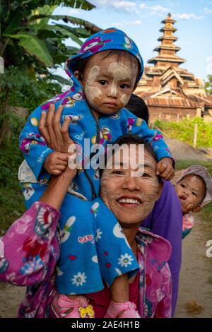 A Burmese mother carries her two infant children down a rural road with their faces powdered with a sunscreen, and with a temple in the background Stock Photo