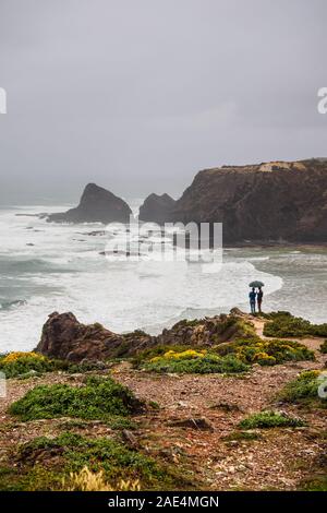 Silhouette of a romantic couple standing on a cliff in the rain under an umbrella, Odeceixe, Algarve, Portugal Stock Photo