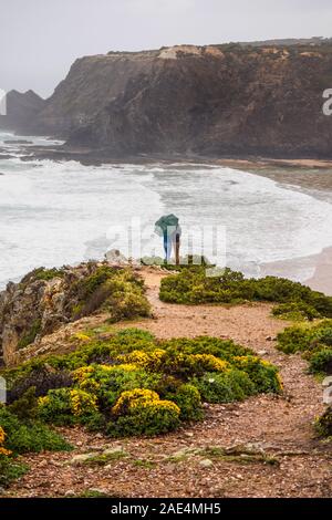 Silhouette of a romantic couple standing on a cliff in the rain under an umbrella, Odeceixe, Algarve, Portugal Stock Photo