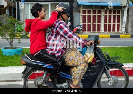 Burmese mother & daughter on a moped with the daughter pushing the mother's helmet too low for her to see on the streets of Mandalay, Myanmar (Burma) Stock Photo