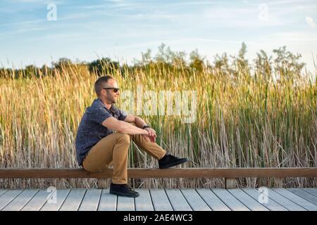 Side view of a peaceful man sitting on boardwalk in park in sunlight Stock Photo