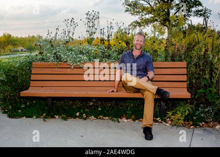A smiling man sits alone on a bench in a beautiful park Stock Photo