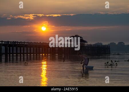 A fisherman stands in shallow water at sunrise in front of the ancient teak wood U Bein Bridge that crosses Taungthaman Lake at Amarapura, near the city of Mandalay in Myanmar (formerly called Burma Stock Photo