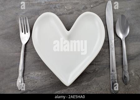White heart shaped plate adorns a rustic dark marble background flanked by detailed silverware. Plenty of copy space on blank plate. Stock Photo