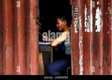 Antipolo City, Philippines – December 5, 2019: Adult Filipino man uses smartphone while inside a warehouse with steel gates. Stock Photo