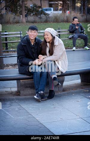 A affectionate Asian couple share a laugh while sitting on a bench in Washington Square Park in Manhattan, New York City. Stock Photo