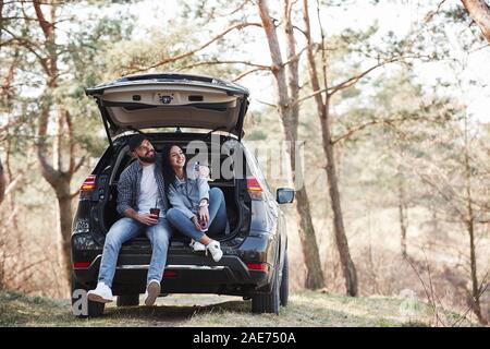 Laughing and having nice time. Sitting on rear part of automobile. Enjoying the nature. Couple have arrived to the forest on their brand new black car Stock Photo