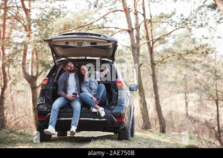 Girl shows next destination. Sitting on rear part of automobile. Enjoying the nature. Couple have arrived to the forest on their brand new black car Stock Photo