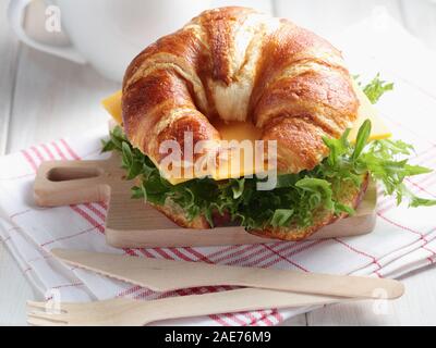 Freshly baked croissant sandwich for quick breakfast  with cheddar cheese, cucumber, and lettuce. Served on small cutting board with wooden fork and k Stock Photo