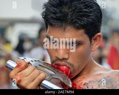 Young Thai Chinese Taoist devotee (mah song) cuts his tongue with a sharp axe during the Phuket Vegetarian Festival (Nine Emperor Gods Festival). Stock Photo