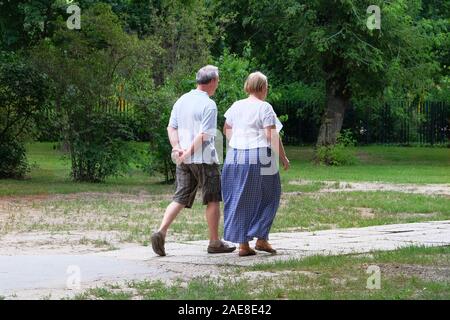 Elderly couple is walking in green city park on day off. Middle aged couple is walking. Green trees on sides of trail. Stock Photo