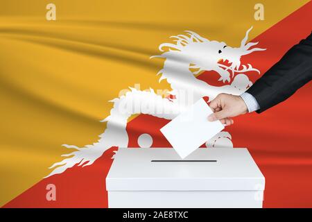 Election in Bhutan. The hand of man putting his vote in the ballot box. Waved Bhutan flag on background. Stock Photo