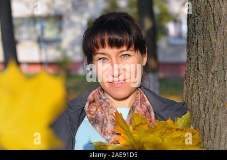 Cute beautiful woman in casual wear holds yellow autumn leaves in her hands on the background of city blocks Stock Photo